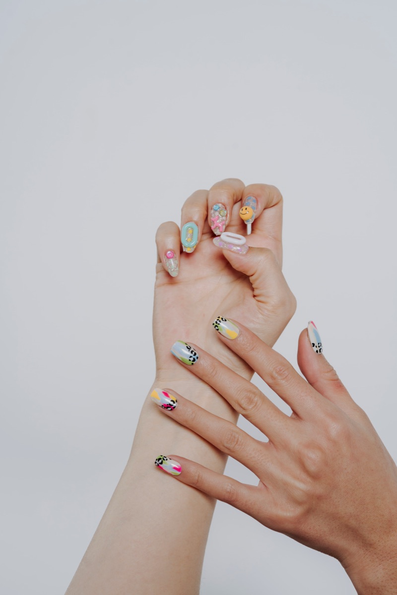 Two hands with long sculpted acrylic nails decorated with elaborate 3D charms — smiley faces, florals, and leopard-print accents
